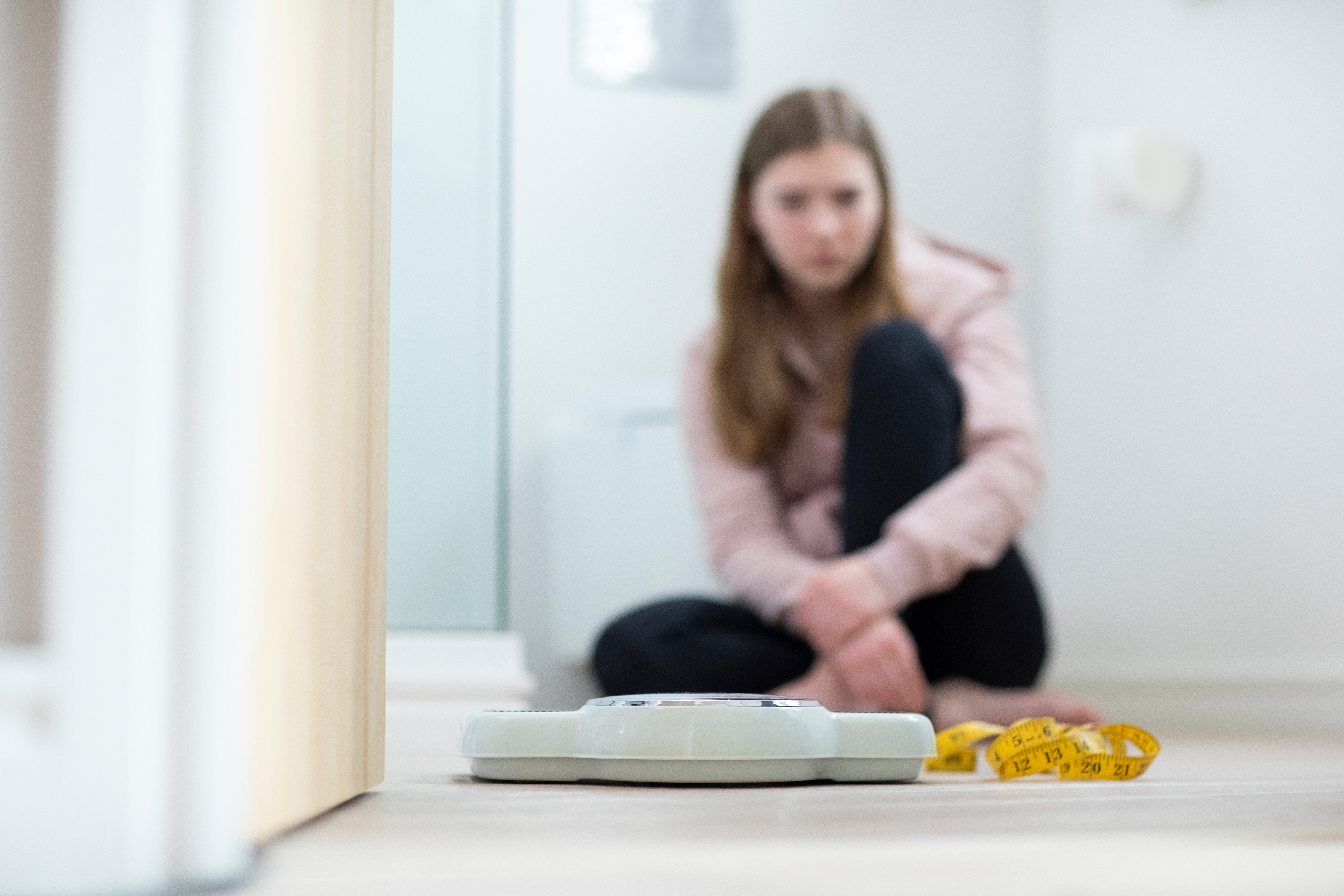 Young girl sits on the bathroom floor staring somberly at a scale and body tape measure, illustrating concern for body image and eating disorders.