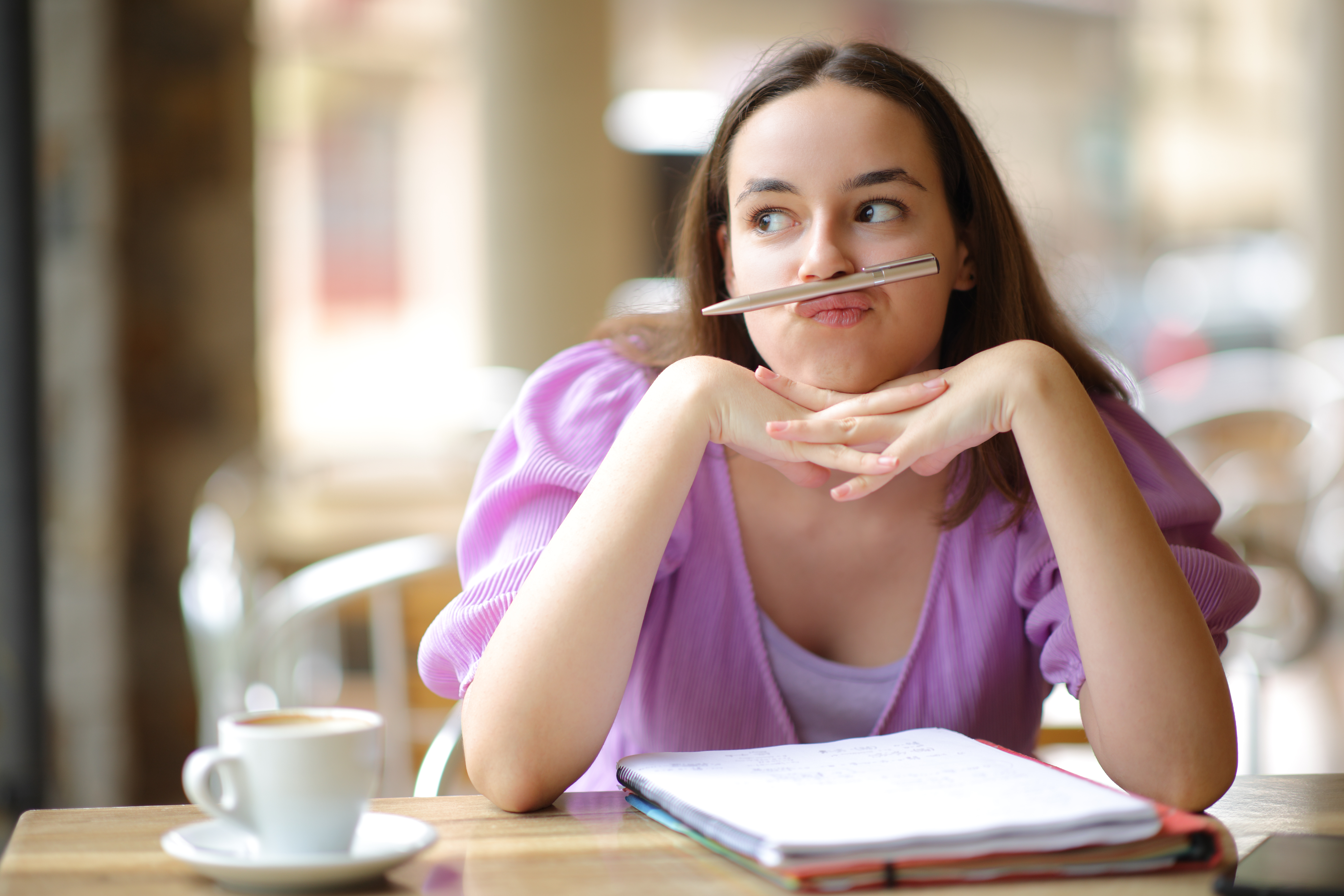 oung woman sits at a desk, with an open notebook in front of her. She sits with her chin on her hands, a pen settled in between her nose and lips, indicating she is distracted and fidgeting to avoid the task in front of her.