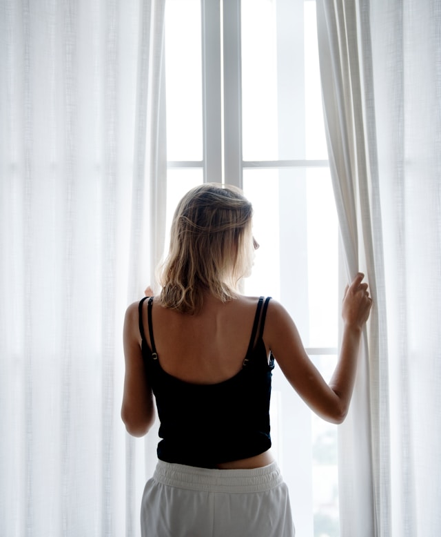 A blonde woman looks out the window, appearing alone and somber.